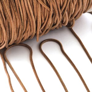 Könnte beinhalten: Eine Spule mit metallisch kupferfarbener Schlangenkette, die eine glatte, flexible und abgerundete Gliederkonstruktion aufweist. Die durchgehende Kette ist fest um einen weißen zylindrischen Kern gewickelt, wobei mehrere lose Stränge herausragen.