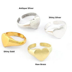 May include: Four adjustable heart-shaped rings in antique silver, shiny silver, shiny gold, and raw brass. Each ring features a smooth heart design and an open, adjustable band. The text labels each ring's finish.