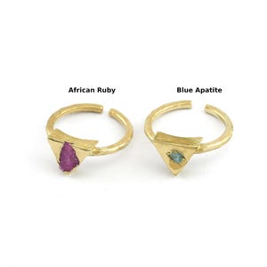 May include: Two gold-colored rings with triangular stone settings. One ring features a vibrant pink African Ruby, and the other showcases a blue apatite stone. The rings have an open design, allowing for size adjustment. The text "African Ruby" and "Blue Apatite" are above each ring.