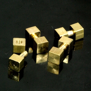 May include: Four gold-colored metal square-shaped pieces with threaded posts. The pieces are arranged in a cluster on a black surface.