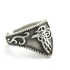 May include: A silver ring with an intricate design featuring a stylized bird or animal. The ring is triangular in shape and has a textured surface.