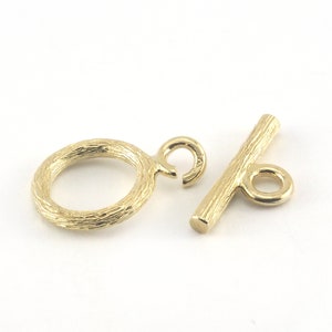 May include: Gold-toned metal toggle clasp with a textured finish. The clasp has a round ring and a bar with a small ring at one end.