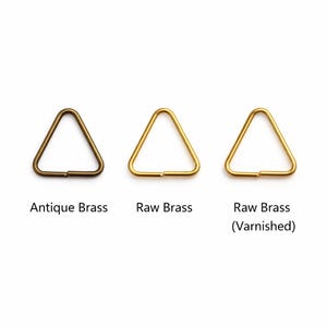 May include: Three triangular metal rings in antique brass, raw brass, and varnished raw brass finishes. Each ring is displayed on a white background, with text labels below each ring indicating the finish.