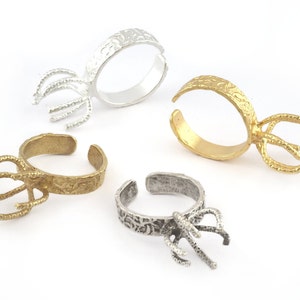 May include: Four adjustable ring blanks with a decorative, textured band and a prong setting. The ring blanks are silver, gold, and two shades of brass.