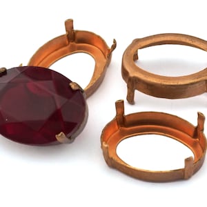May include: Three gold-toned metal settings for oval-shaped gemstones. One setting has a red glass gemstone in place.