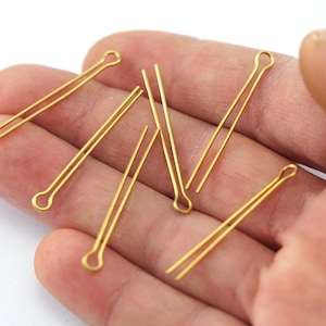 Op de afbeelding: Zes goudkleurige kopspelden met een lus aan het ene uiteinde. De spelden zijn waaiervormig op een hand gerangschikt.