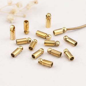 May include: A selection of small, gold-coloured metal jewellery findings. The findings are cylindrical with a rounded end and a small ball-shaped detail. Some are lying down, others stand upright.