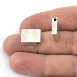 Puede incluir: Dos cuentas metálicas rectangulares plateadas con acabado mate. Cada cuenta tiene un pequeño agujero perforado. Una cuenta está plana, la otra está de pie, mostrando el agujero. Estas cuentas son probablemente para hacer joyas.
