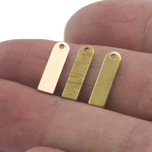 Puede incluir: Tres dijes de metal en forma rectangular, en diferentes acabados: oro, latón y oro rosa. Cada dije es rectangular con un pequeño agujero en la parte superior para sujetarlo a un collar o pulsera.