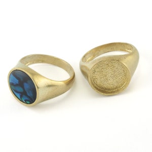 May include: Two gold signet rings. One ring has a blue and green abalone shell inlay. The other ring has a smooth, blank surface.