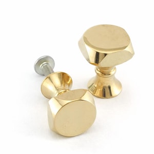 Puede incluir: Dos tiradores dorados para armarios con diseño geométrico. Cada tirador tiene una parte superior hexagonal facetada y una base decorativa. Un tirador está unido a un tornillo plateado. Los tiradores son probablemente para muebles o decoración.