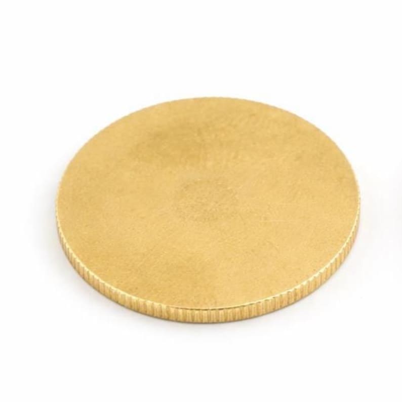 Op de afbeelding: Een ronde, goudkleurige metalen schijf met een getextureerde rand. De schijf heeft een eenvoudig ontwerp, mogelijk een munt of een decoratief element. Het oppervlak heeft een subtiele, matte afwerking. Diameter ongeveer 5 cm.