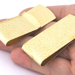 Puede incluir: Dos sujetabilletes dorados con un acabado texturizado y martillado. Un clip está abierto, revelando una superficie lisa y plana. El otro clip está cerrado. Los clips se sostienen en una mano.