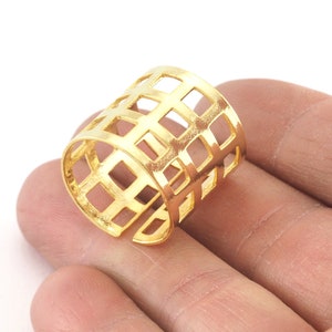 May include: A gold-colored ring with a geometric, openwork design. The ring features a series of square cutouts, creating a cage-like appearance. The ring is adjustable and sits on a finger, showcasing its bright, metallic finish.