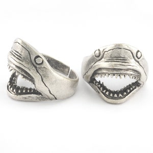 May include: Two silver rings with a shark head design. The rings are open and have a detailed shark head with an open mouth and sharp teeth.