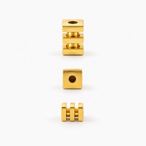 May include: Three gold-colored metal beads are displayed against a white background. One bead is a stack of three square blocks. Another is a single square block. The third bead has three vertical bars.