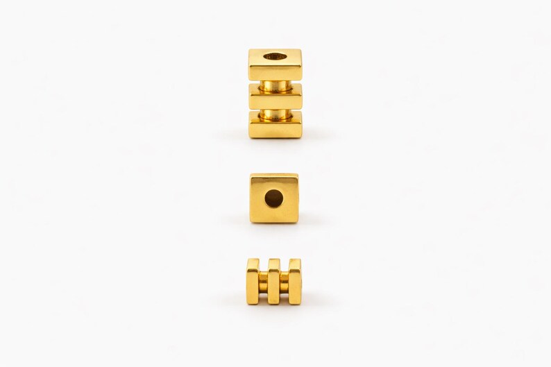 May include: Three gold-colored metal beads are displayed against a white background. One bead is a stack of three square blocks. Another is a single square block. The third bead has three vertical bars.