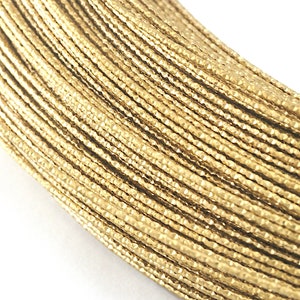 May include: Close-up of a bundle of gold-colored wire, tightly packed together. The wire has a textured, faceted surface, catching the light and creating a shimmering effect. The background is plain white, highlighting the metallic sheen of the wire.