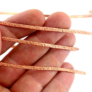 May include: A long, thin, flexible strip of copper metal featuring a distinct, uneven, hammered texture across its entire surface. The copper wire is coiled in a spiral shape, showcasing its metallic reddish-brown color and reflective finish.