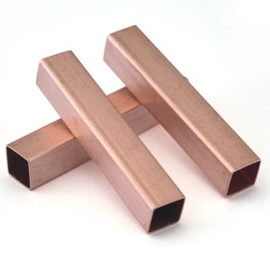 Pode incluir: Três tubos retangulares de cor cobre são dispostos sobre uma superfície branca. Os tubos têm uma seção transversal quadrada e são abertos em ambas as extremidades. O metal tem uma superfície ligeiramente refletora.