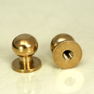 May include: Two gold-toned metal knobs with a round top and a threaded base. The knobs are designed for furniture or other objects.