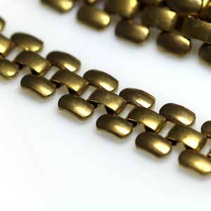 Puede incluir: Una cadena de metal dorado con un diseño de eslabones. La cadena está hecha de pequeños eslabones rectangulares que están conectados entre sí.