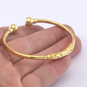 May include: A gold-colored open cuff bracelet. The bracelet has a decorative center section and ball-shaped ends. The bracelet is shown against a light background.