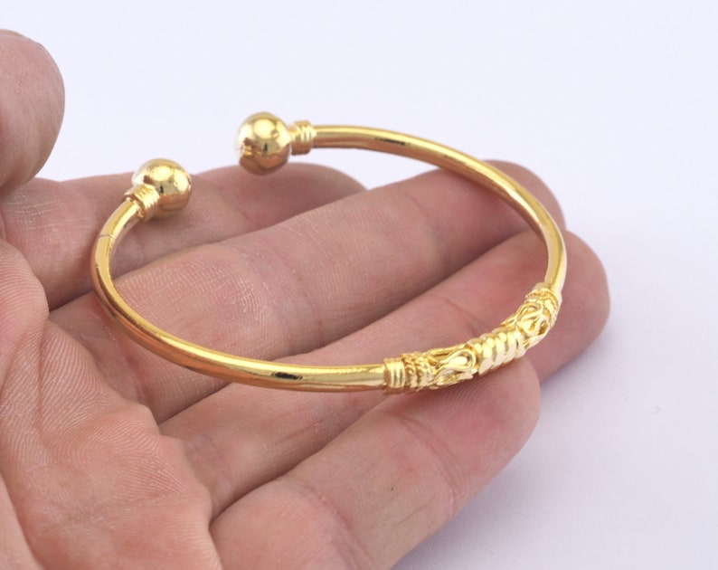 May include: A gold-colored open cuff bracelet. The bracelet has a decorative center section and ball-shaped ends. The bracelet is shown against a light background.