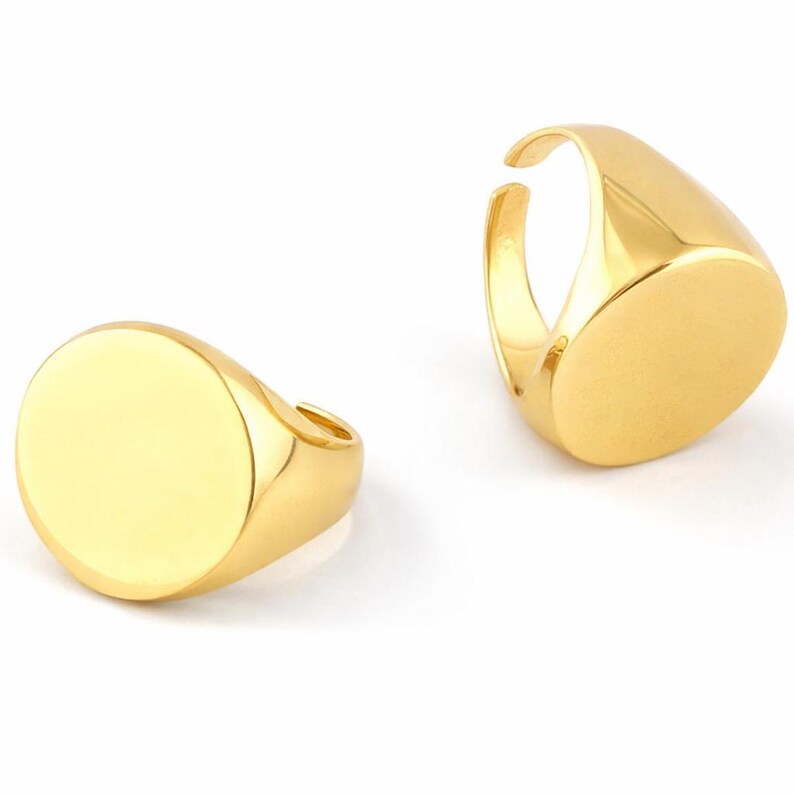 May include: Two gold-colored signet rings. Each ring has a smooth, oval face and a thick band. The rings are displayed against a white background, showcasing their polished, reflective surfaces. The design is simple and classic.