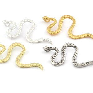 Puede incluir: Una colección de colgantes con forma de serpiente en varios acabados metálicos. Los colgantes son de plata, oro y un color plata envejecida más oscuro. Cada serpiente tiene un patrón texturizado similar a escamas.