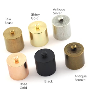 Puede incluir: Colección de tapones de botella cilíndricos de metal en varios acabados: latón crudo, oro brillante, plata antigua, oro rosa, negro y bronce antiguo. Cada tapón tiene una pequeña parte superior redonda. Las etiquetas identifican cada acabado.