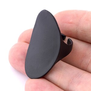 Peut inclure: Une bague noire mate avec une face ovale allongée. La bague est tenue dans une main sur fond blanc. La bague est réglable.