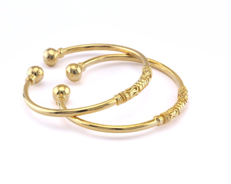 May include: Two gold-colored bangles with ball-shaped ends and decorative detailing. The bangles are displayed on a white background. One bangle has a detailed pattern along its band. The other bangle has a smooth, simple design.