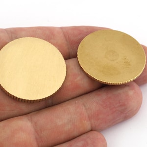 Op de afbeelding: Twee ronde, goudkleurige metalen schijven. Elke schijf heeft een glad, plat midden en een geribbelde rand. De schijven worden in een hand tegen een witte achtergrond gehouden. De schijven lijken van messing of een vergelijkbaar metaal te zijn.