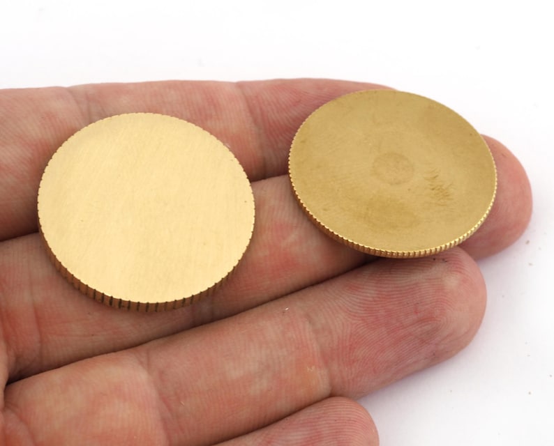 Op de afbeelding: Twee ronde, goudkleurige metalen schijven. Elke schijf heeft een glad, plat midden en een geribbelde rand. De schijven worden in een hand tegen een witte achtergrond gehouden. De schijven lijken van messing of een vergelijkbaar metaal te zijn.