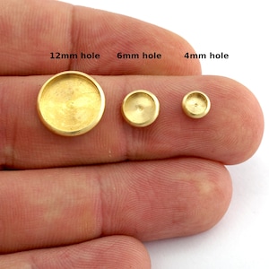 May include: Three small, gold-colored metal bezels of varying sizes are displayed. The largest bezel has a 12mm hole, the middle one has a 6mm hole, and the smallest has a 4mm hole. The bezels are held in a person's hand.