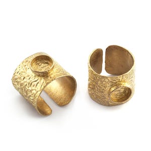 May include: Two gold-toned open rings featuring a wide, textured band with a hammered finish. Each ring includes a single circular bezel setting on the outer surface, designed to hold a stone or cabochon. The metal has a matte, brushed appearance.