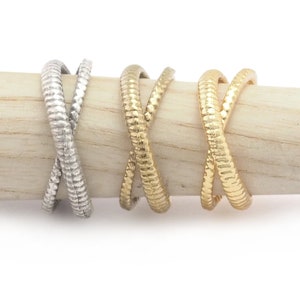 May include: Three textured metal rings in silver and gold tones. The rings have a crisscross design, with a snake-like texture. The rings are displayed on a light-colored wooden dowel against a white background.