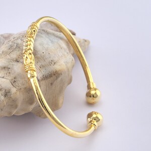 May include: A gold-colored cuff bracelet with decorative detailing. The bracelet has a curved design with a decorative section in the middle and ball-shaped ends. The bracelet is displayed on a seashell against a white background.