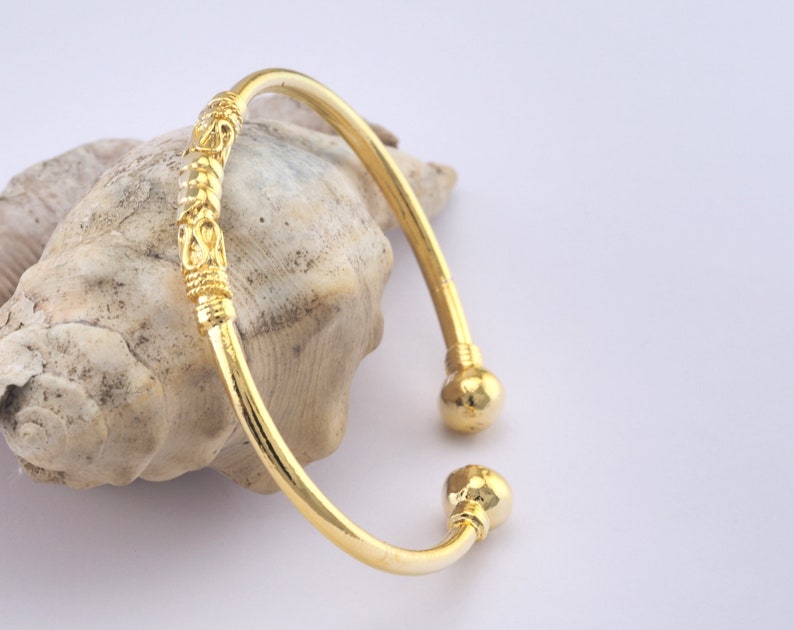 May include: A gold-colored cuff bracelet with decorative detailing. The bracelet has a curved design with a decorative section in the middle and ball-shaped ends. The bracelet is displayed on a seashell against a white background.
