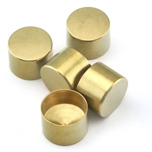 May include: A collection of five small, cylindrical brass objects. Each has a flat top and bottom with a smooth, polished finish. The objects are arranged on a white surface, with varying orientations, showcasing their simple, geometric design.