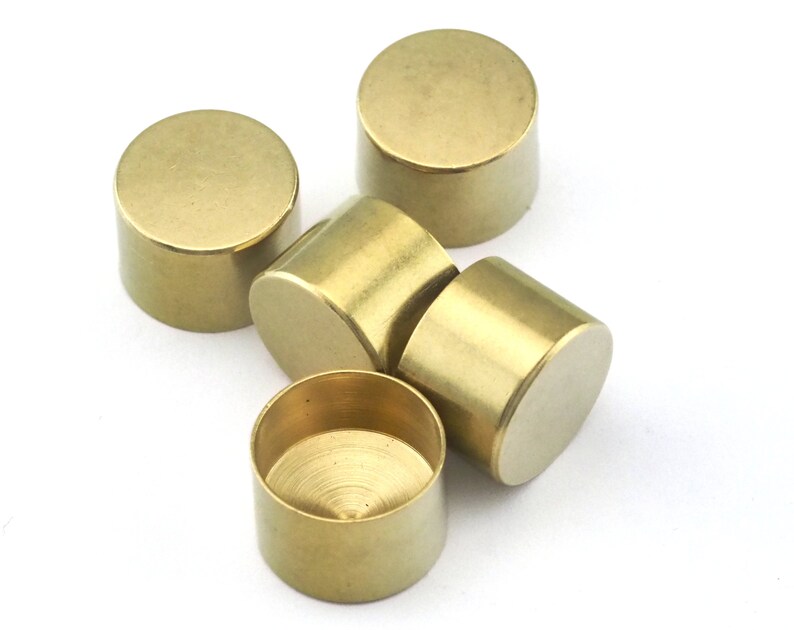 May include: A collection of five small, cylindrical brass objects. Each has a flat top and bottom with a smooth, polished finish. The objects are arranged on a white surface, with varying orientations, showcasing their simple, geometric design.