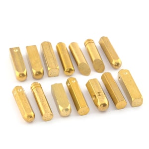 May include: A collection of 15 gold-colored metal stamp heads, each with a unique shape and design. The stamp heads are arranged in two rows on a white surface.