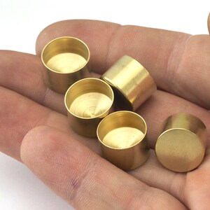 May include: A collection of small, cylindrical brass components with a rounded base and open top. The components are displayed in a hand, showcasing their metallic sheen and simple design. These components are likely for jewelry making or other craft projects.