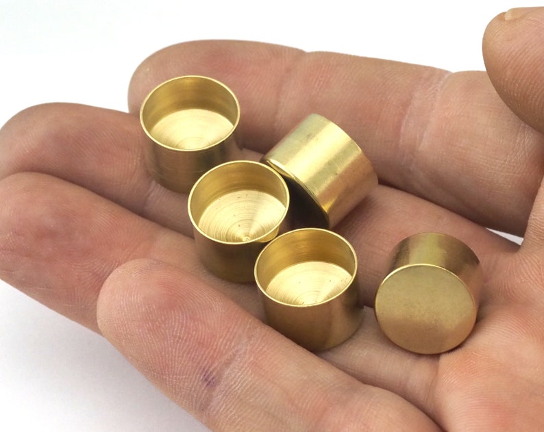 May include: A collection of small, cylindrical brass components with a rounded base and open top. The components are displayed in a hand, showcasing their metallic sheen and simple design. These components are likely for jewelry making or other craft projects.