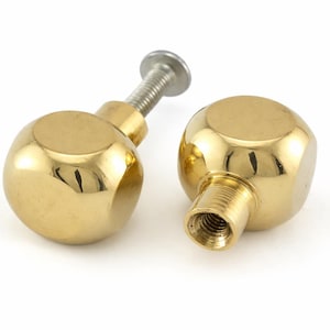May include: Two polished brass cabinet knobs with a spherical design. One knob has a threaded post and a silver screw, while the other has a threaded hole. The knobs are on a white background.