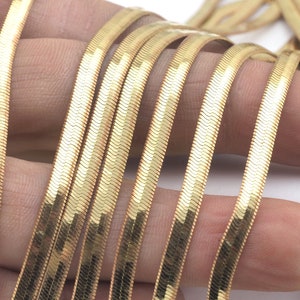 以下が含まれることがあります： 金色のヘビの鎖のネックレスのクローズアップ。チェーンは、滑らかで流れるようなデザインを生み出す小さな連鎖するリンクでできています。