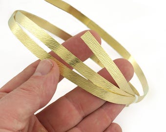 May include: A hand holds a coil of textured, gold-colored metal strips. The strips have a subtle, linear pattern and are arranged in a circular shape. The background is plain white, highlighting the metallic sheen of the material.