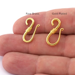 May include: Two gold-coloured hook clasps, one labelled "Raw Brass" and the other "Gold Plated". The clasps have a curved hook shape with a decorative knot detail. The clasps are displayed against a neutral background.