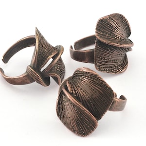 Może przedstawiać: Trzy pierścionki w kolorze miedzi o unikalnych, teksturowanych wzorach. Pierścionki mają regulowaną obrączkę i charakteryzują się abstrakcyjnym, warstwowym kształtem. Pierścionki są prezentowane na białym tle, ukazując ich skomplikowane detale i metaliczne wykończenie.